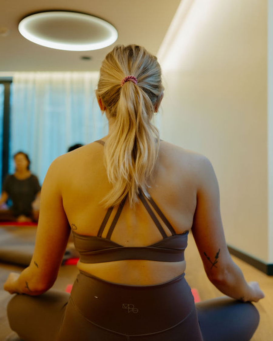 Blonde Frau im Yogasitz im Vordergrund, umgeben von weiteren Teilnehmern in einer Yoga-Session