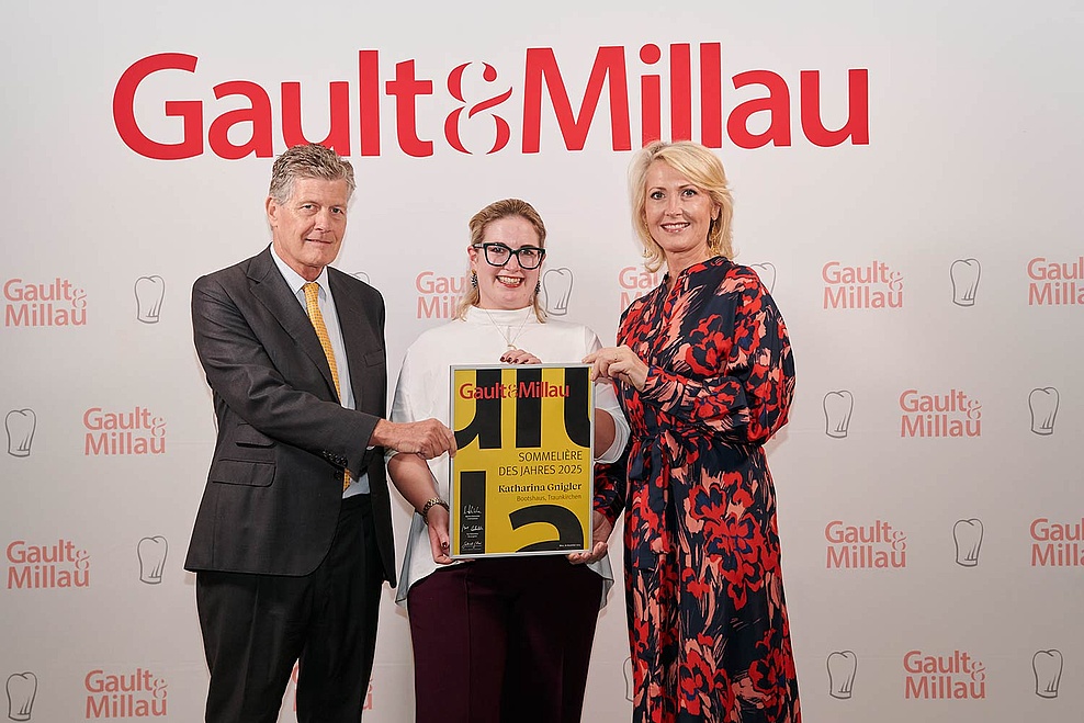 Katharina, Gewinnerin der Gault&Millau Auszeichnung für Sommeliere des Jahres 2025, steht mit ihrer Auszeichnung in der Mitte. Neben ihr stehen ein Mann und eine Dame, die die Auszeichnung überreichen, vor einem großen weißen Plakat mit roter Schrift.