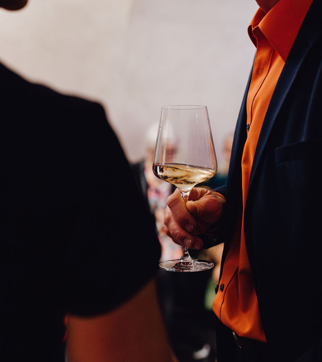 Mann in blauem Anzug mit orangefarbenem Hemd hält ein Glas Weißwein, während zwei Personen im schwarzen T-Shirt nur von hinten sichtbar sind.