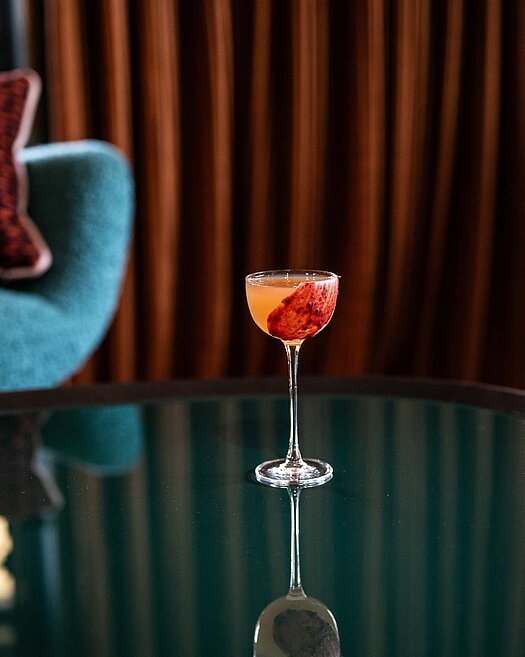 Orangefarbener Cocktail auf einem Tisch, im Hintergrund ein dunkler Vorhang