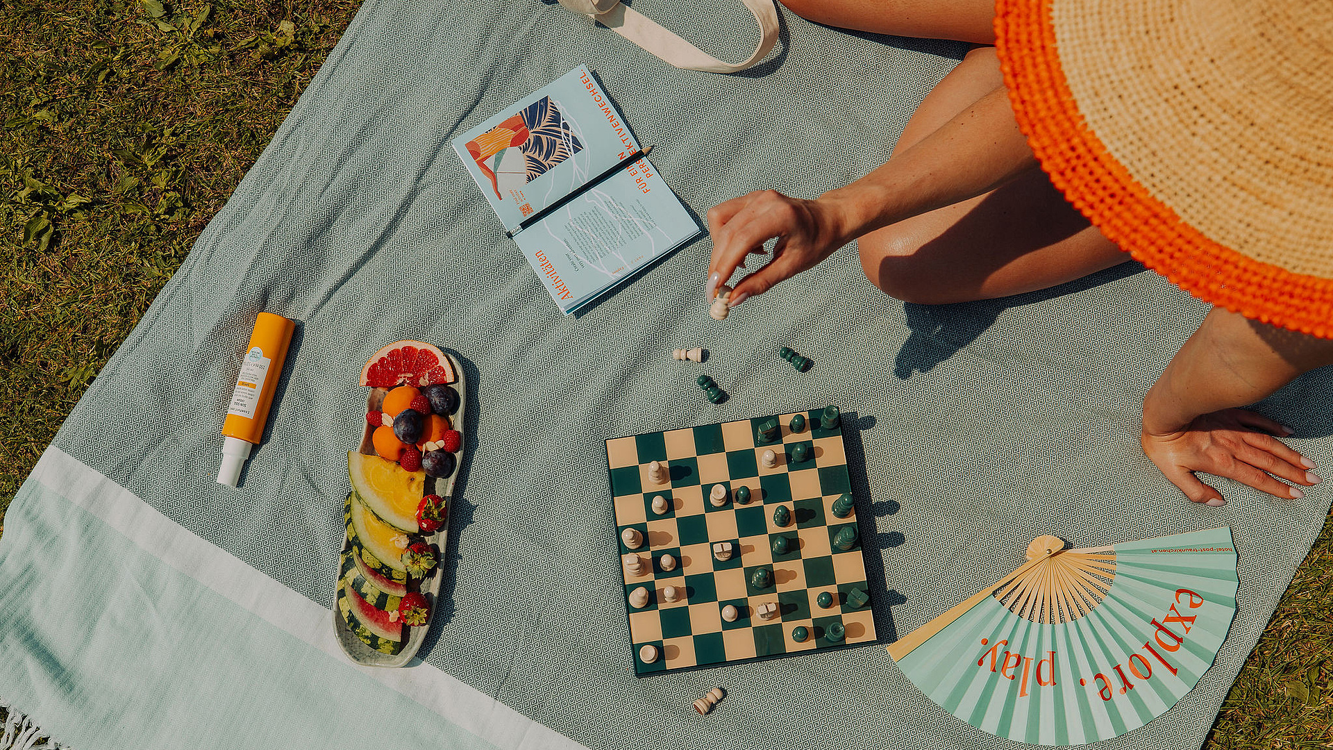 Frau mit großem Strohhut sitzt auf grauer Decke auf der Wiese und spielt Schach. Um sie herum liegen ein Fächer, eine Obstplatte, Sonnencreme und ein Buch.