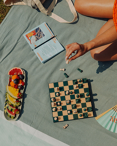 Frau mit großem Strohhut sitzt auf grauer Decke auf der Wiese und spielt Schach. Um sie herum liegen ein Fächer, eine Obstplatte, Sonnencreme und ein Buch.