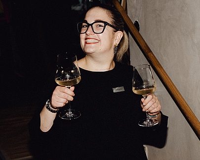 Sommelière im schwarzen Kleid lächelt mit zwei Weingläsern in der Hand, im Restaurant aufgenommen