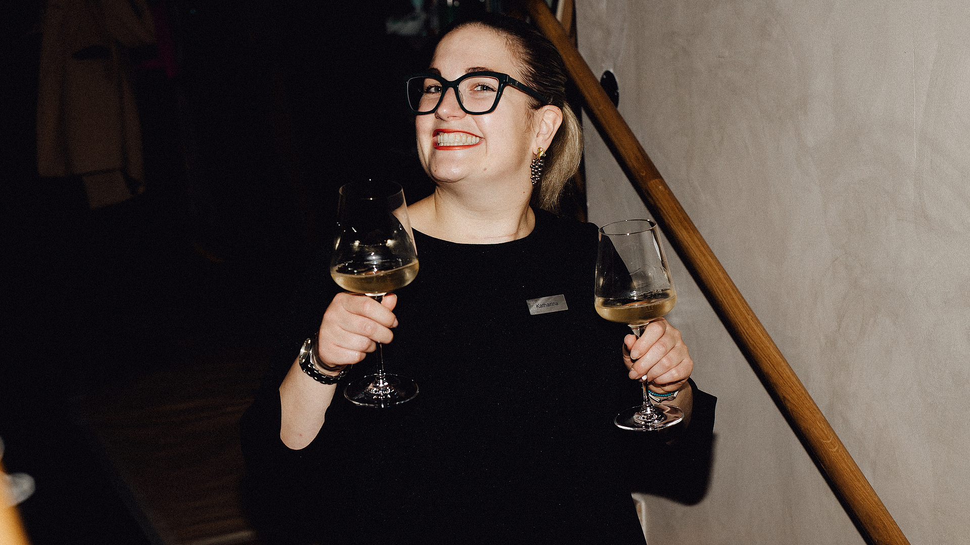 Sommelière im schwarzen Kleid lächelt mit zwei Weingläsern in der Hand, im Restaurant aufgenommen