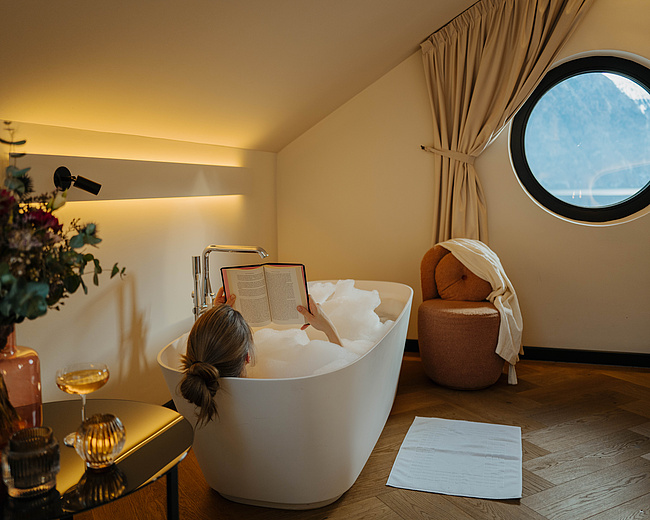 Dreamy Lake Junior Suite in der Post am See mit freistehender Badewanne, rundem Fenster und stilvollem Badezimmer.