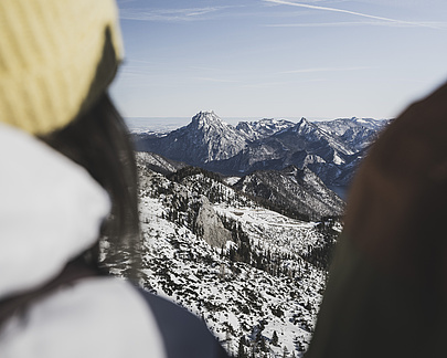 Frau in Skijacke und Mütze von hinten, die die schneebedeckte Berglandschaft im Winter betrachtet.