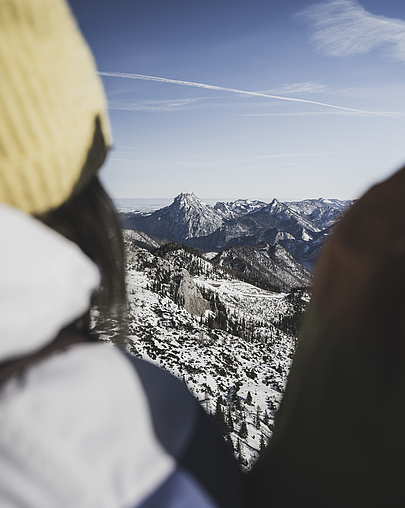 Frau in Skijacke und Mütze von hinten, die die schneebedeckte Berglandschaft im Winter betrachtet.