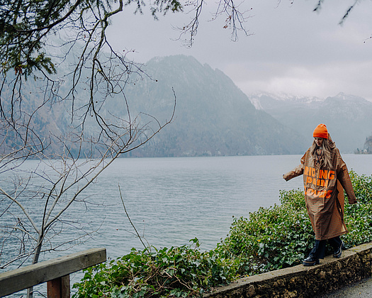 Person mit orangefarbener Mütze und braunem Regencape balanciert im Regen auf einer Steinmauer, im Hintergrund die Berge und der Traunsee