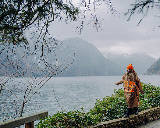 Person mit orangefarbener Mütze und braunem Regencape balanciert im Regen auf einer Steinmauer, im Hintergrund die Berge und der Traunsee