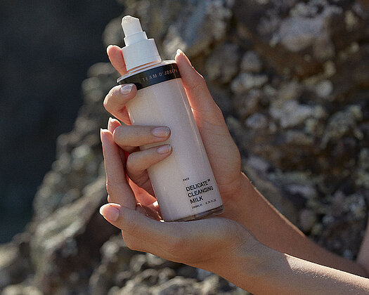 Beige Flasche "Delicate Cleansing Milk" von Team Dr. Sommer wird in Händen gehalten, mit Steinen und Berg im Hintergrund, Sonne und Schatten.