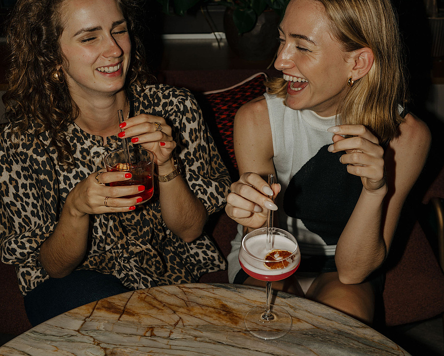 Zwei Frauen sitzen an einem runden Tisch im Barbereich und genießen Cocktails, während sie lachen