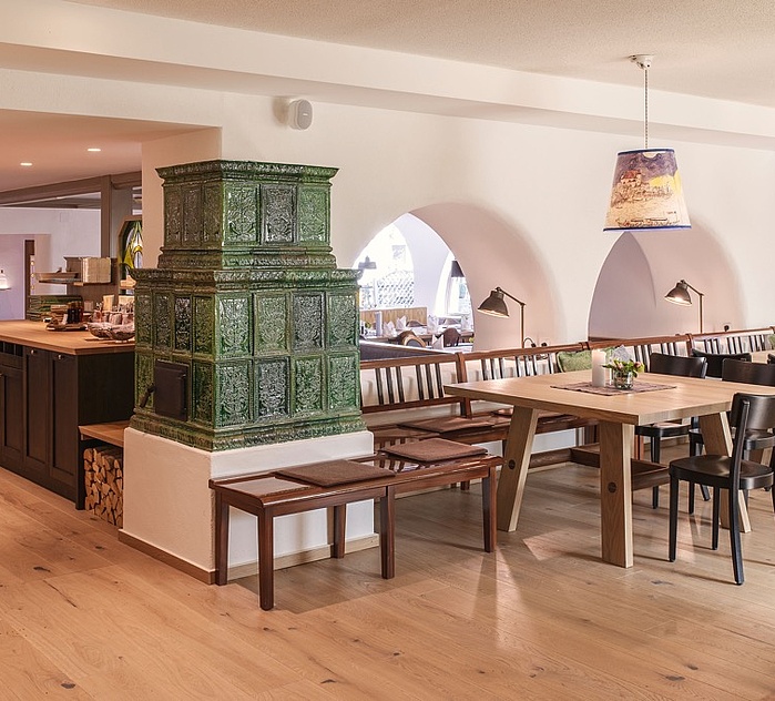 Innenansicht des Restaurants Poststube mit großen, hellen Holztischen, einem Kachelofen mit grünen Fliesen und hellem Holzboden