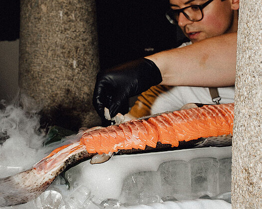 Koch beim Anrichten eines großen Fisches auf Eiswürfeln mit Rauch, auf einer weißen Tischdecke.