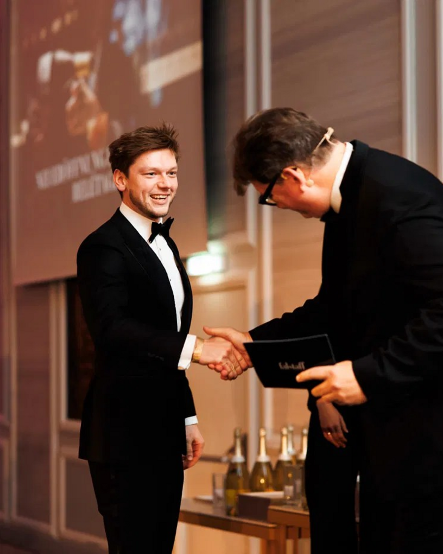 Markus bei der Übergabe der Falstaff-Auszeichnung, während er einem Vertreter die Hand schüttelt, beide lächeln in einer festlichen Umgebung.