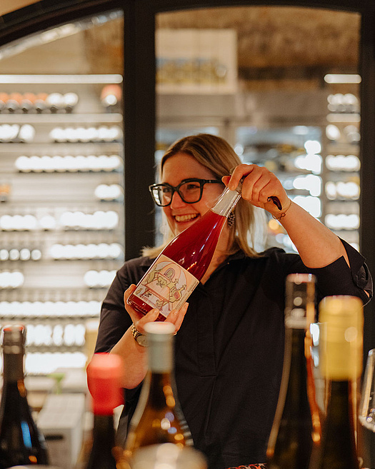 Person schenkt hinter der Bar Wein aus einer Flasche in ein schräg gehaltenes Weinglas, im Hintergrund beleuchtete Weinregale.