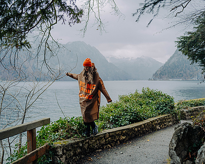 Person im braunen Regencape balanciert auf einer Steinmauer, im Hintergrund die Berge und der Traunsee