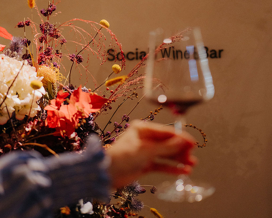 Hand mit Weinglas im Vordergrund, dahinter ein buntes Blumenarrangement und der Schriftzug „Social Wine Bar“ an der Wand