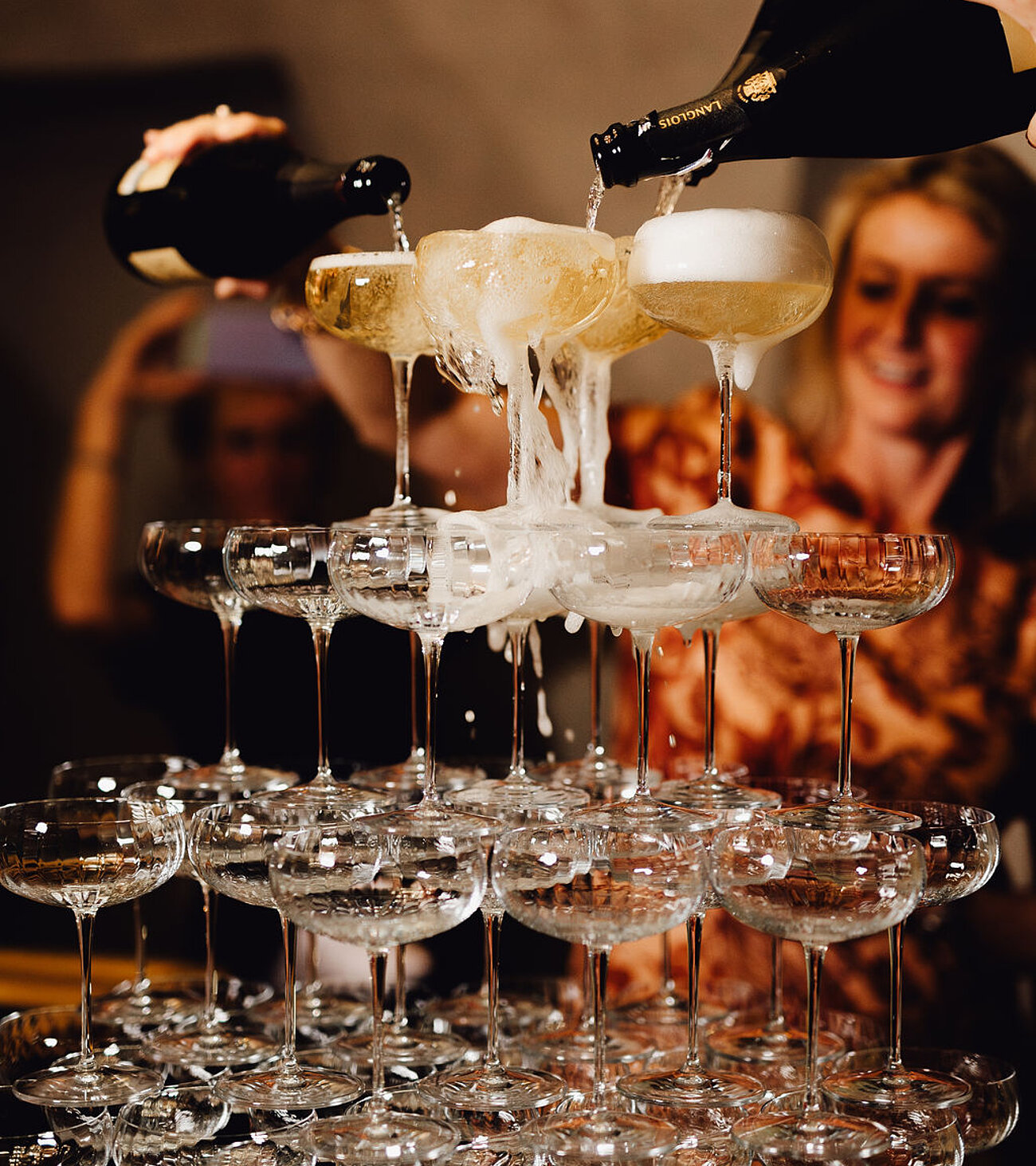 Gläserturm mit Champagner, der von oben eingegossen wird, im Hintergrund Partygäste.
