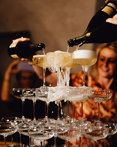 Gläserturm mit Champagner, der von oben eingegossen wird, im Hintergrund Partygäste.