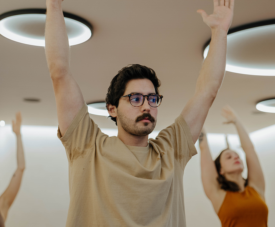 Mann mit Brille praktiziert Yoga im Vordergrund, während zwei Frauen im Hintergrund ebenfalls Übungen durchführen
