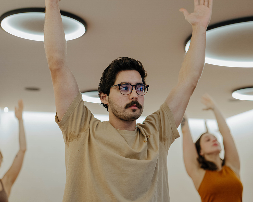 Mann mit Brille praktiziert Yoga im Vordergrund, während zwei Frauen im Hintergrund ebenfalls Übungen durchführen