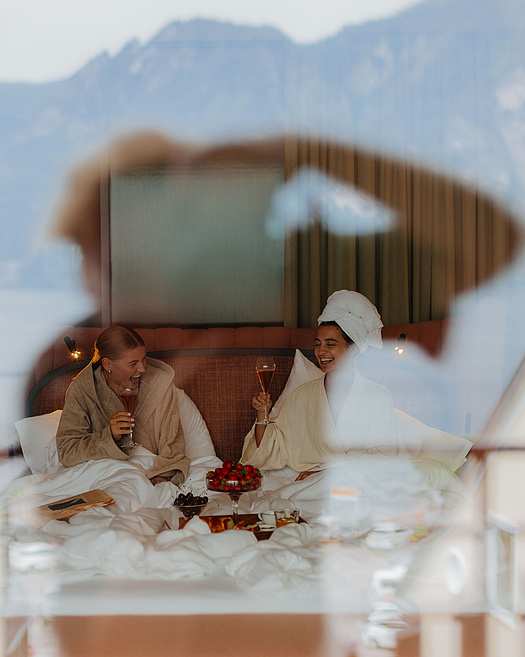 Zwei Frauen im Bademantel sitzen lachend im Bett mit einem Glas Sekt in der Hand. Vor ihnen steht ein Tablett mit süßen Köstlichkeiten. In der Glasreflexion sind der Fotograf, der See und die Berge zu sehen.