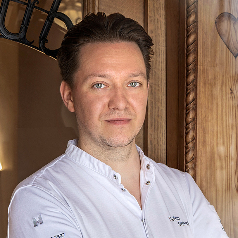 Koch in weißer Kochjacke am Eingang des Restaurants vor einer Holztür.