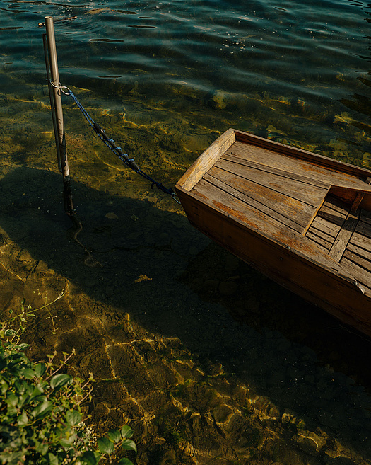 Holzboot (Plätte) angebunden an Metallpfahl im Wasser, ruhige Atmosphäre.