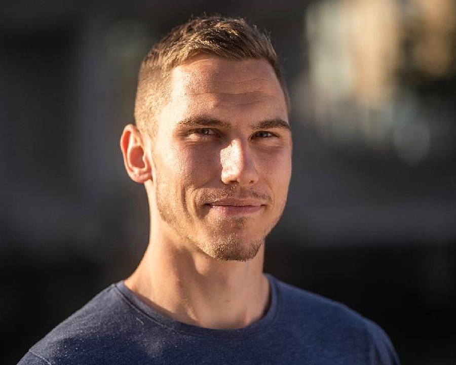 Mann im blauen Pullover mit kurzem Haar und Bart, dessen Gesicht von der Sonne beleuchtet wird.
