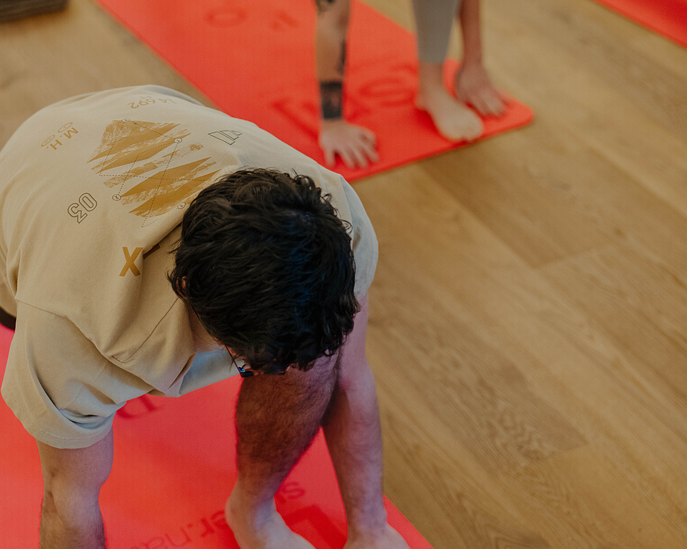 Zwei Teilnehmer in Yoga-Posen auf einer roten Matte im Yogaraum mit Holzboden