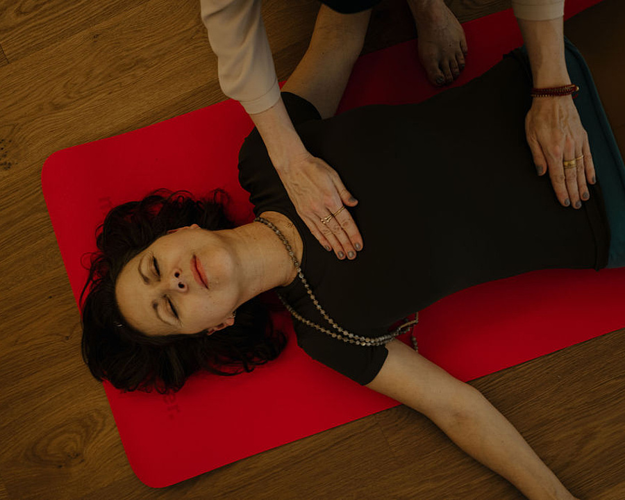 Teilnehmerin bei einer Breathwork-Session liegt entspannt auf einer roten Yogamatte, während die Hände der Kursleiterin sanft auf ihrem Bauch ruhen