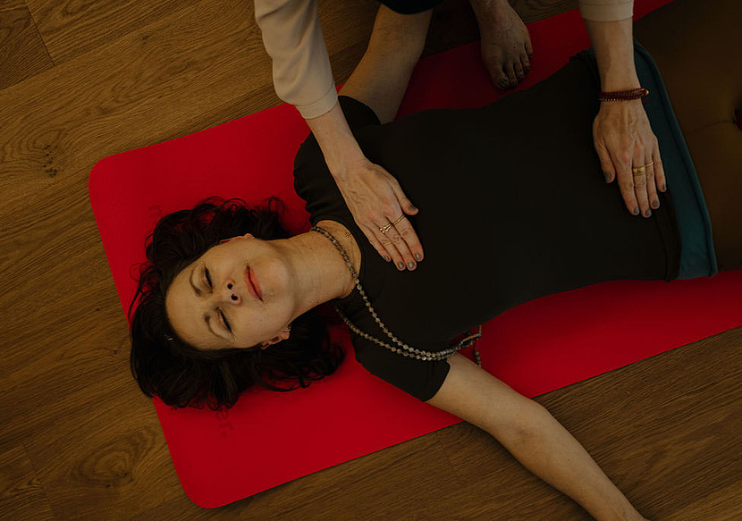 Teilnehmerin bei einer Breathwork-Session liegt entspannt auf einer roten Yogamatte, während die Hände der Kursleiterin sanft auf ihrem Bauch ruhen