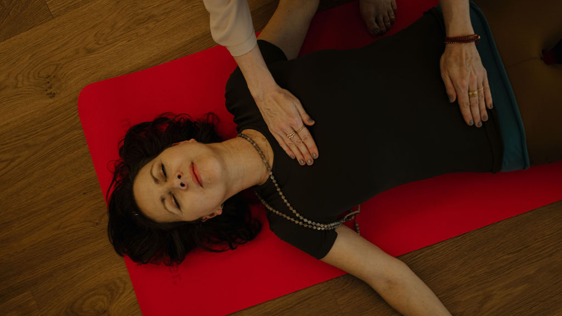 Teilnehmerin bei einer Breathwork-Session liegt entspannt auf einer roten Yogamatte, während die Hände der Kursleiterin sanft auf ihrem Bauch ruhen