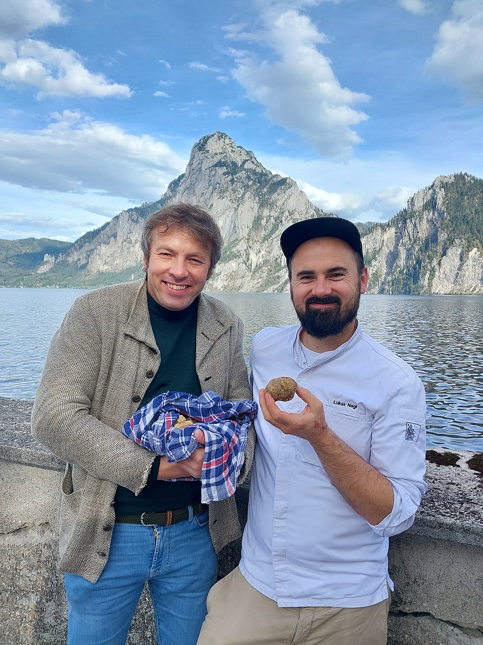 Küchenchef und Trüffelsammler halten lächelnd eine Trüffel in der Hand, mit Traunsee und Traunstein im Hintergrund.