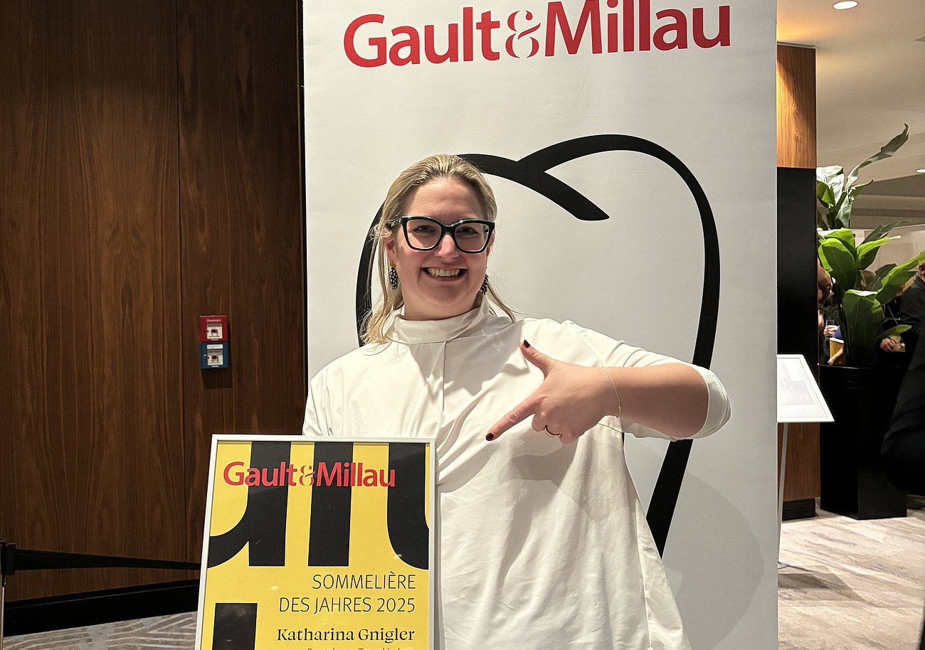 Katharina hält stolz ihre Auszeichnung in der Hand und steht vor einem Gault&Millau Banner in der Lobby. Sie lächelt und strahlt Freude über ihren Erfolg aus.