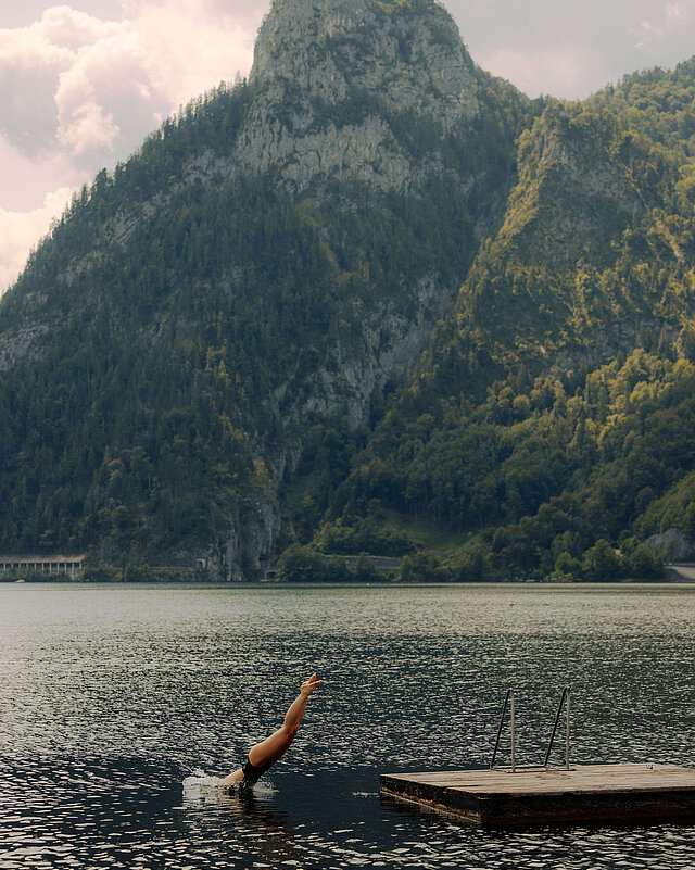 Person springt ins Wasser des Traunsees von einer Holzinsel aus, im Hintergrund Berge und ein klarer Sommerhimmel.