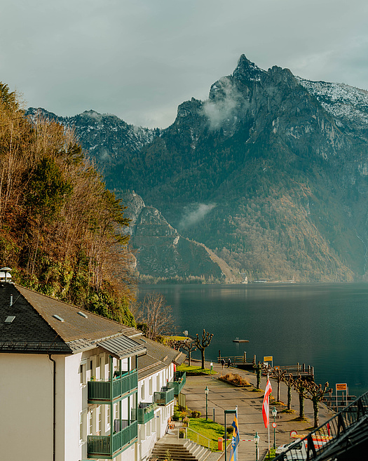 Traunsee mit der „Schlafenden Griechin“ im Hintergrund, weißes Haus mit Flaggen, Holzstege am Ufer.