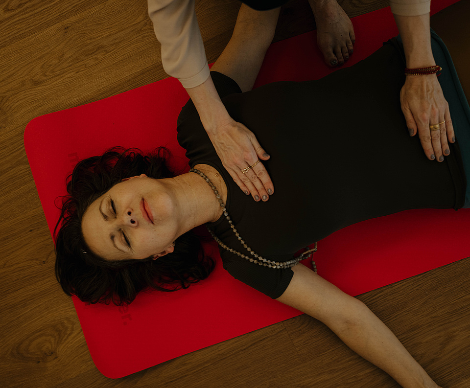 Teilnehmerin bei einer Breathwork-Session liegt entspannt auf einer roten Yogamatte, während die Hände der Kursleiterin sanft auf ihrem Bauch ruhen.