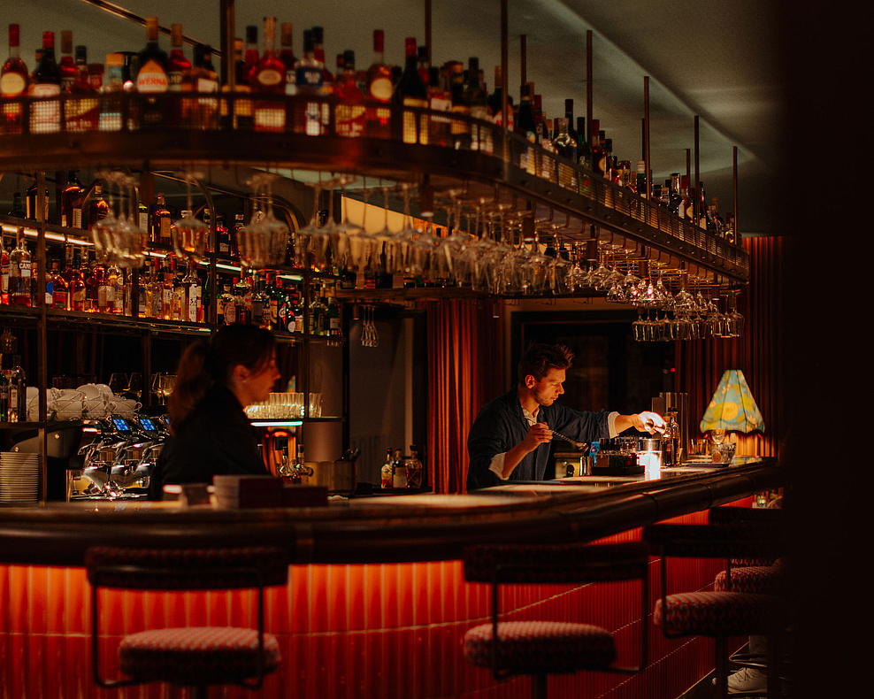 Der Barbereich mit gemütlichen Barstühlen, einer gut bestückten Bar und einem Barkeeper, der Getränke zubereitet