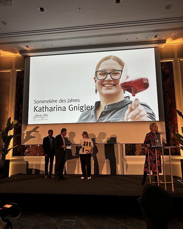 Katharina nimmt die Auszeichnung als Sommeliere des Jahres entgegen, während ein großes Foto von ihr mit Weinglas auf einer Leinwand im Hintergrund zu sehen ist. Vertretern stehen neben ihr und gratulieren ihr.