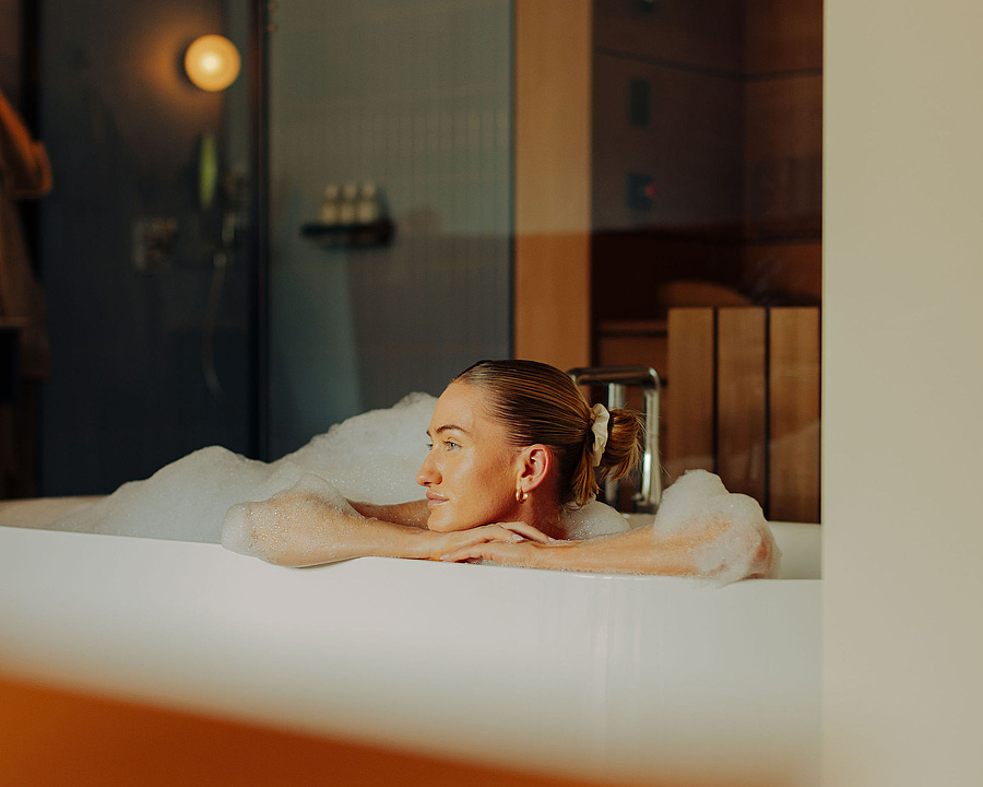 Frau mit zusammengebundenen Haaren entspannt in einer Schaumbadewanne, den Kopf auf ihre Arme gestützt. Im Hintergrund sind eine Dusche und eine Sauna zu sehen.