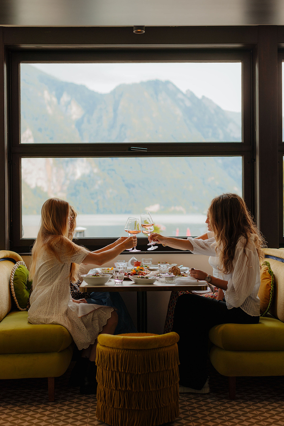 Vier Frauen genießen ein gemeinsames Abendessen, stoßen an und teilen sich Speisen, während im Hintergrund die Berge und der See sichtbar sind