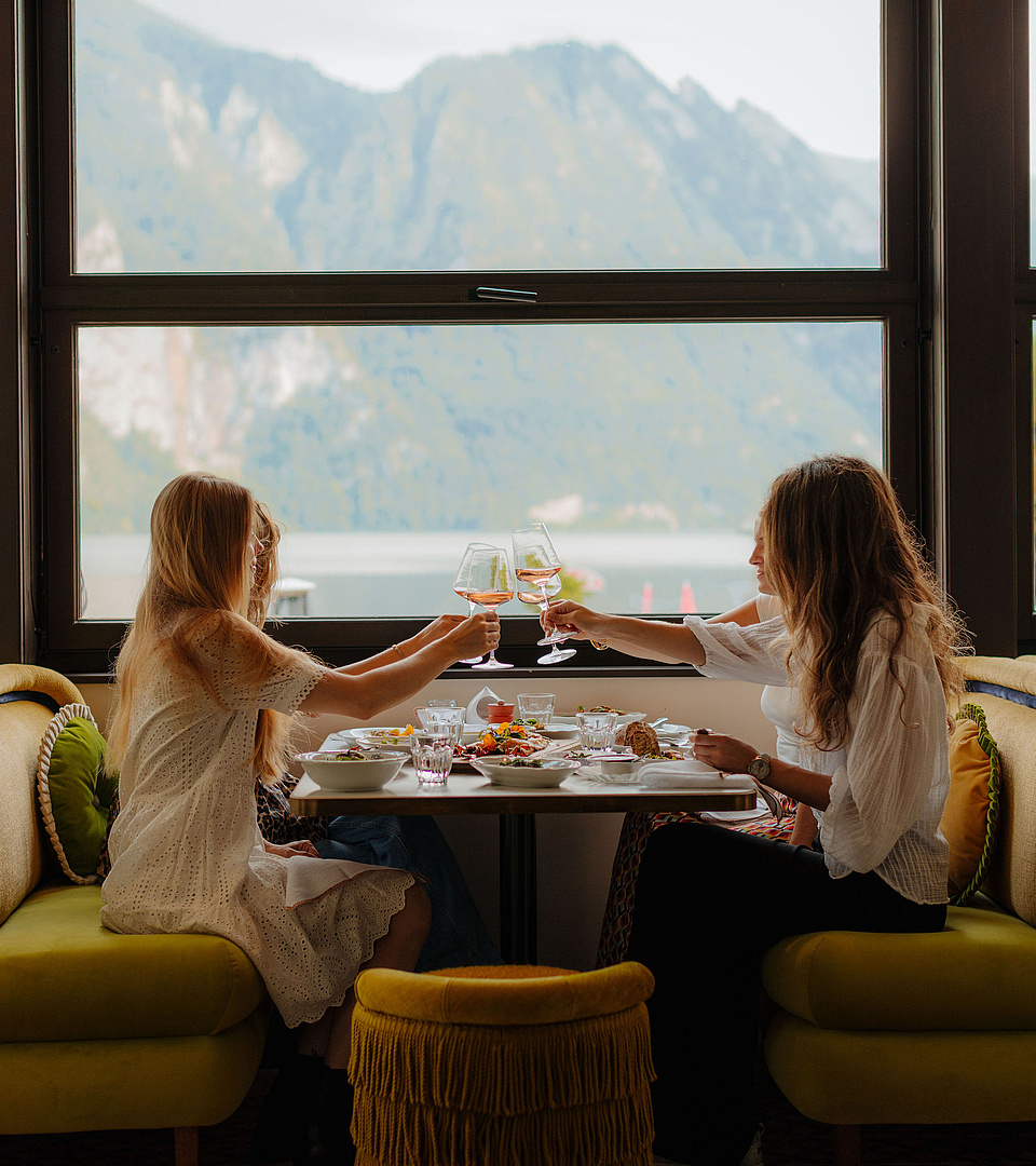 Vier Frauen genießen ein gemeinsames Abendessen, stoßen an und teilen sich Speisen, während im Hintergrund die Berge und der See sichtbar sind