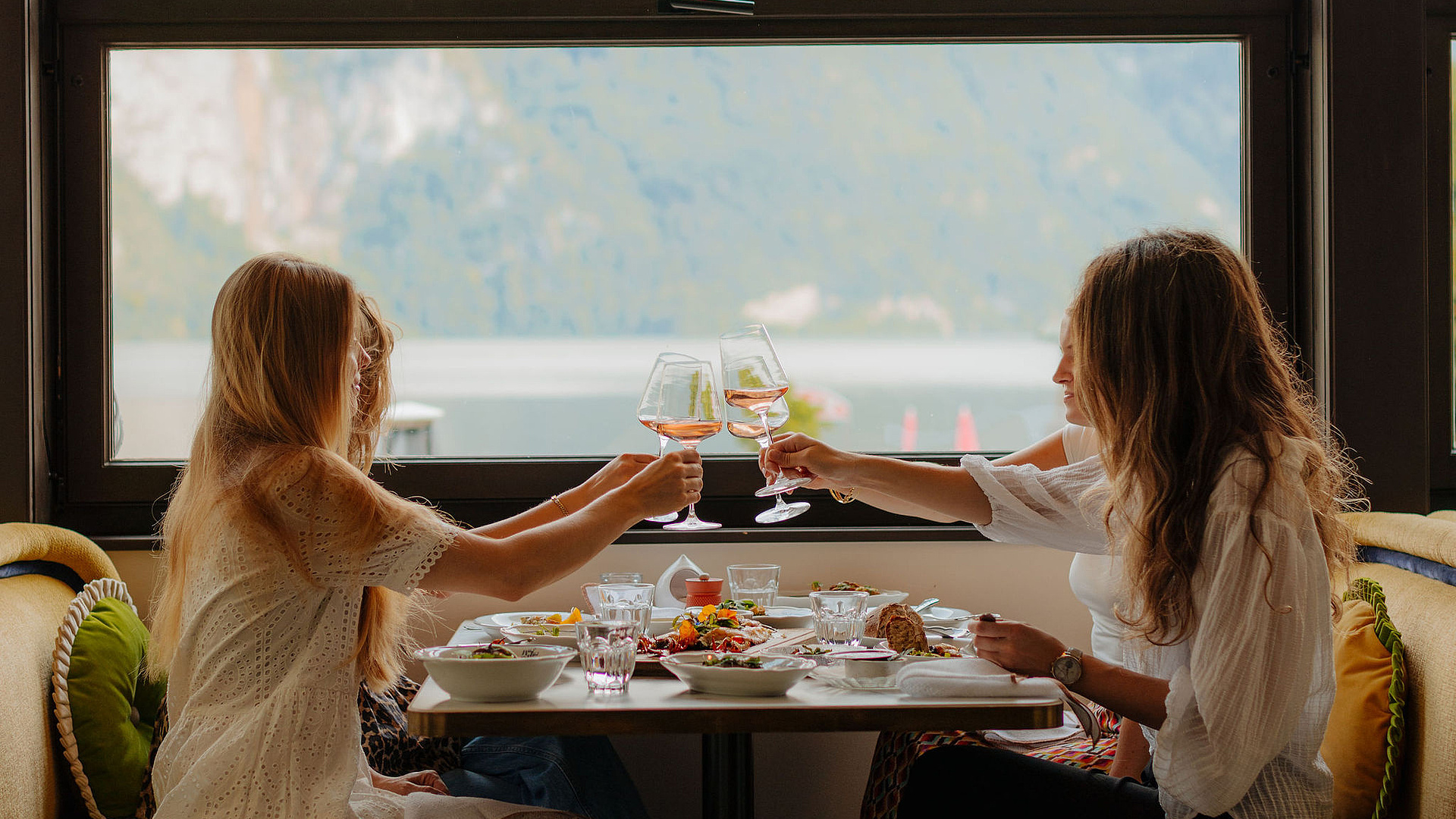 Vier Frauen genießen ein gemeinsames Abendessen, stoßen an und teilen sich Speisen, während im Hintergrund die Berge und der See sichtbar sind