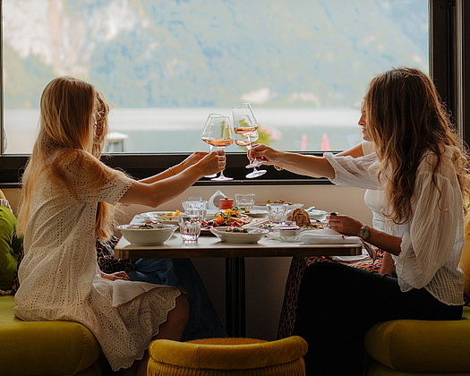 Vier Frauen genießen ein gemeinsames Abendessen, stoßen an und teilen sich Speisen, während im Hintergrund die Berge und der See sichtbar sind