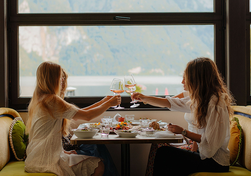 Vier Frauen genießen ein gemeinsames Abendessen, stoßen an und teilen sich Speisen, während im Hintergrund die Berge und der See sichtbar sind