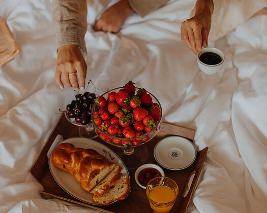 Zwei Frauen im Bett genießen ihren Morgenkaffee. Eine von ihnen nimmt sich eine Kirsche von einem liebevoll angerichteten Tablett mit frischen Früchten und süßen Köstlichkeiten.