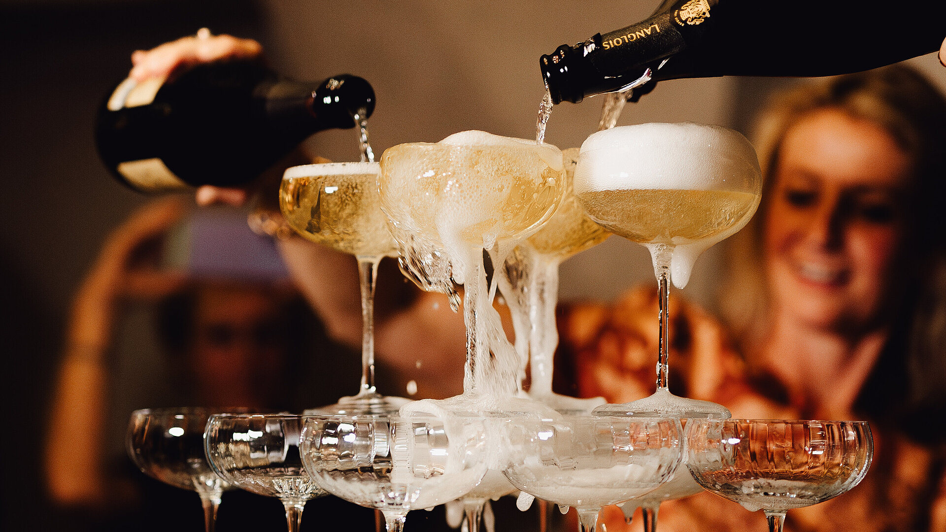 Gläserturm mit Champagner, der von oben eingegossen wird, im Hintergrund Partygäste.