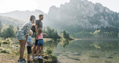 Eine Familie steht am Seeufer des Laudachsee in Grünberg im Salzkammergut und schaut aufs Wasser, die Berge spiegeln sich im See, (c) Oberösterreich Tourismus/Traunsee-Almtal/Robert Maybach