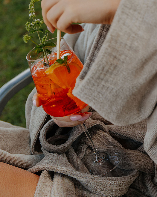 Frau im Bademantel sitzt draußen auf einem Stuhl in der Wiese und hält einen orangen Cocktail in der Hand.